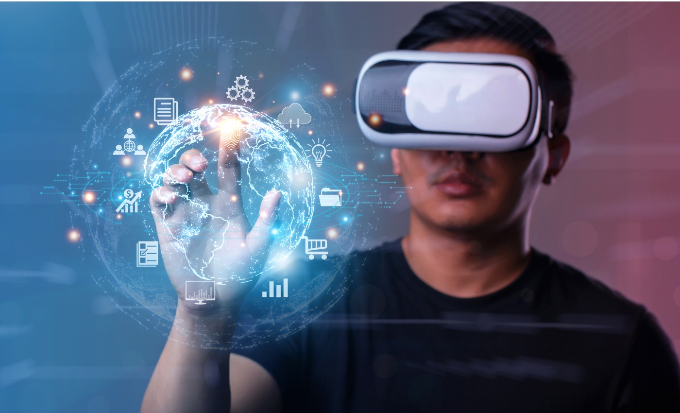 A clean-shaven man in a blue t-shirt wearing a VR headset holds his finger up to a hologram of financial icons circling the globe in this image demonstrating fintech in the Metaverse.