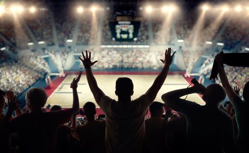 Cheering basketball fans are silhouetted over the court from the grandstands in this image representing sports analytics.