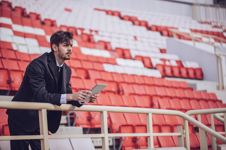 A sports executive stands amidst empty red and white bleachers, holding a tablet with sports analytics data and looking out at the team.