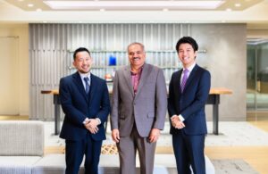 Three professionally dressed men standing in a modern office lobby, smiling at the camera.