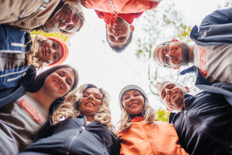 Group of people forming a circle, looking down at the camera and smiling outdoors.