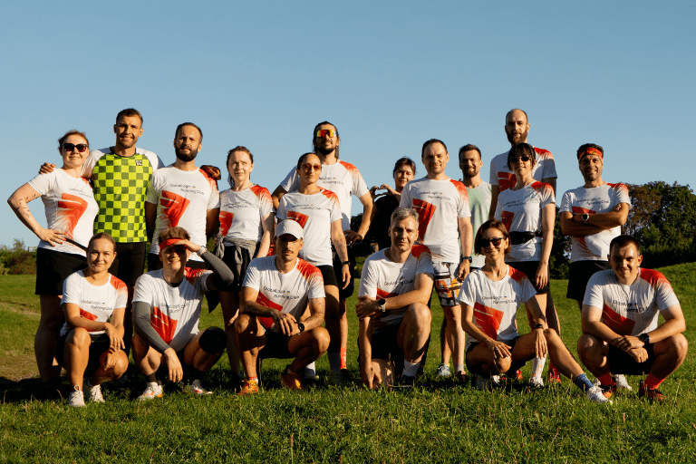 Group of runners in matching white shirts with red accents posing on a sunny grassy field. The team stands and kneels together under a clear blue sky.
