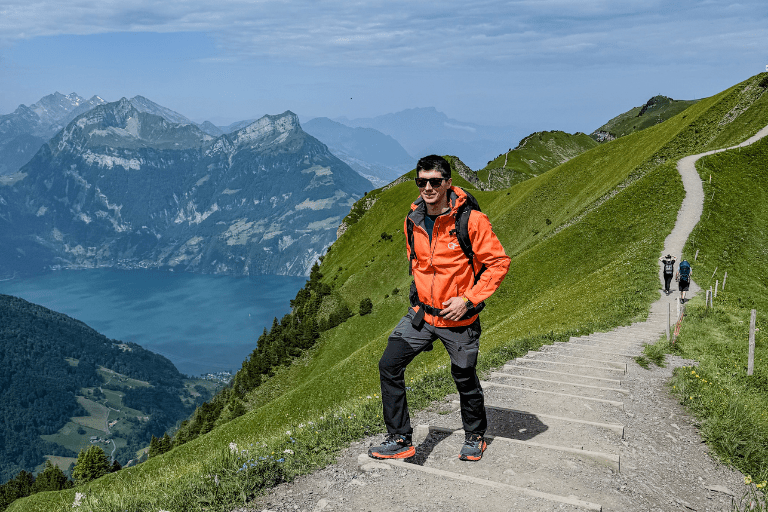 Man in an orange jacket and sunglasses stands on a mountain trail with green slopes and steps, overlooking a blue lake and distant peaks