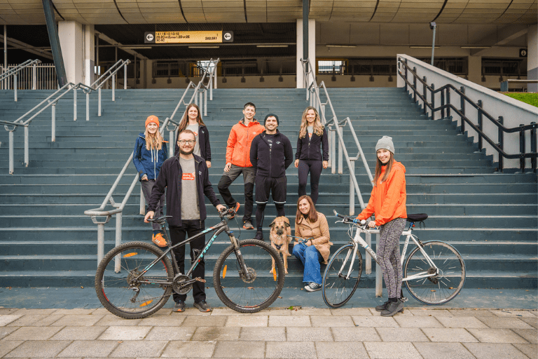 Eight people posing on a wide outdoor staircase with two bicycles and a dog in front of a building.
