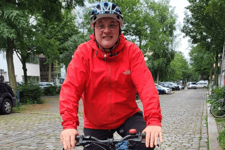Person wearing a red jacket and helmet riding a bicycle toward the camera on a tree-lined city street.