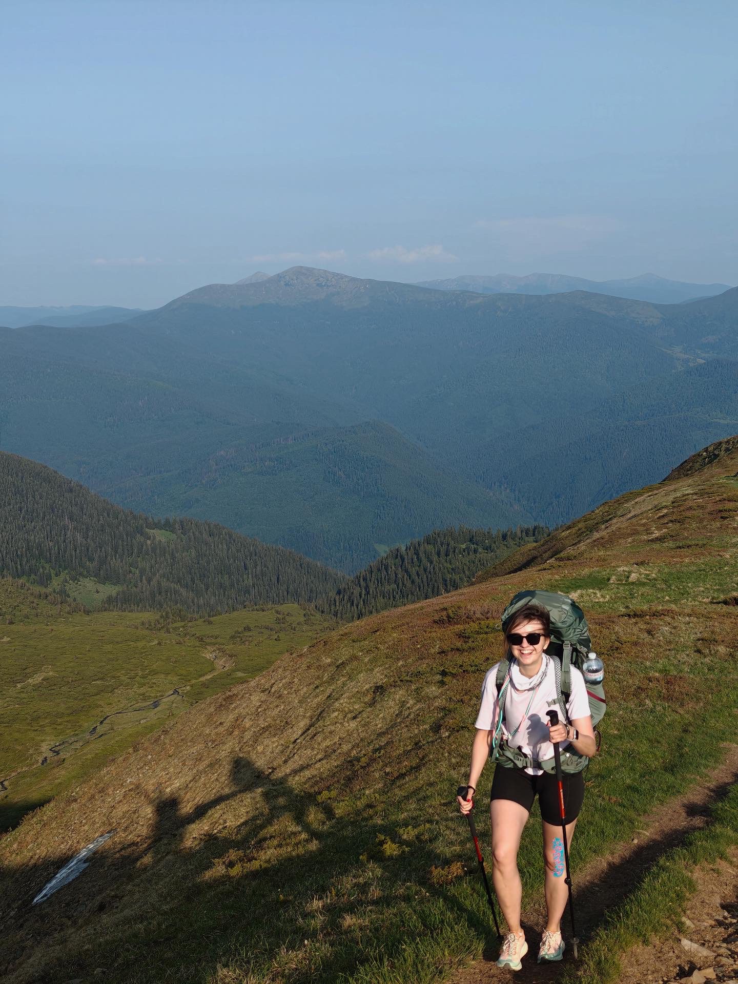 Smiling female hiker with a large backpack and trekking poles on a hillside trail, with green valleys and distant mountains in the background.