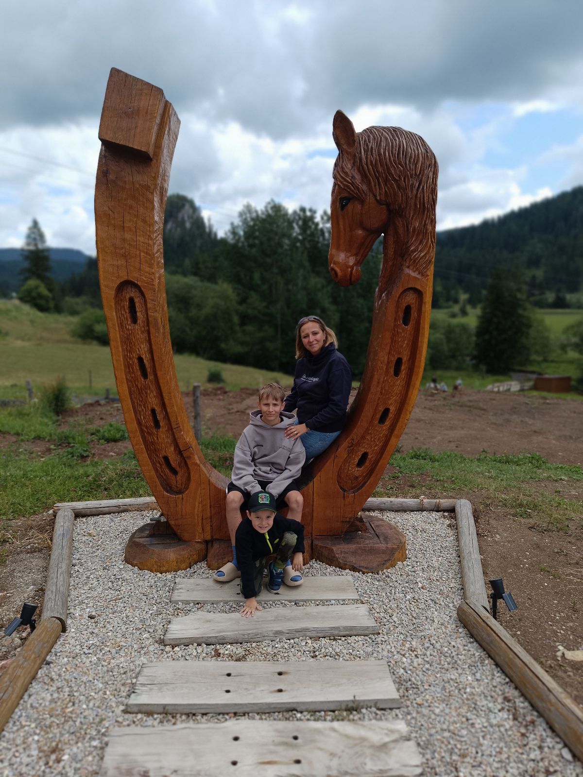 Family posing on a large carved wooden horse sculpture outdoors, with hills and cloudy sky in the background.