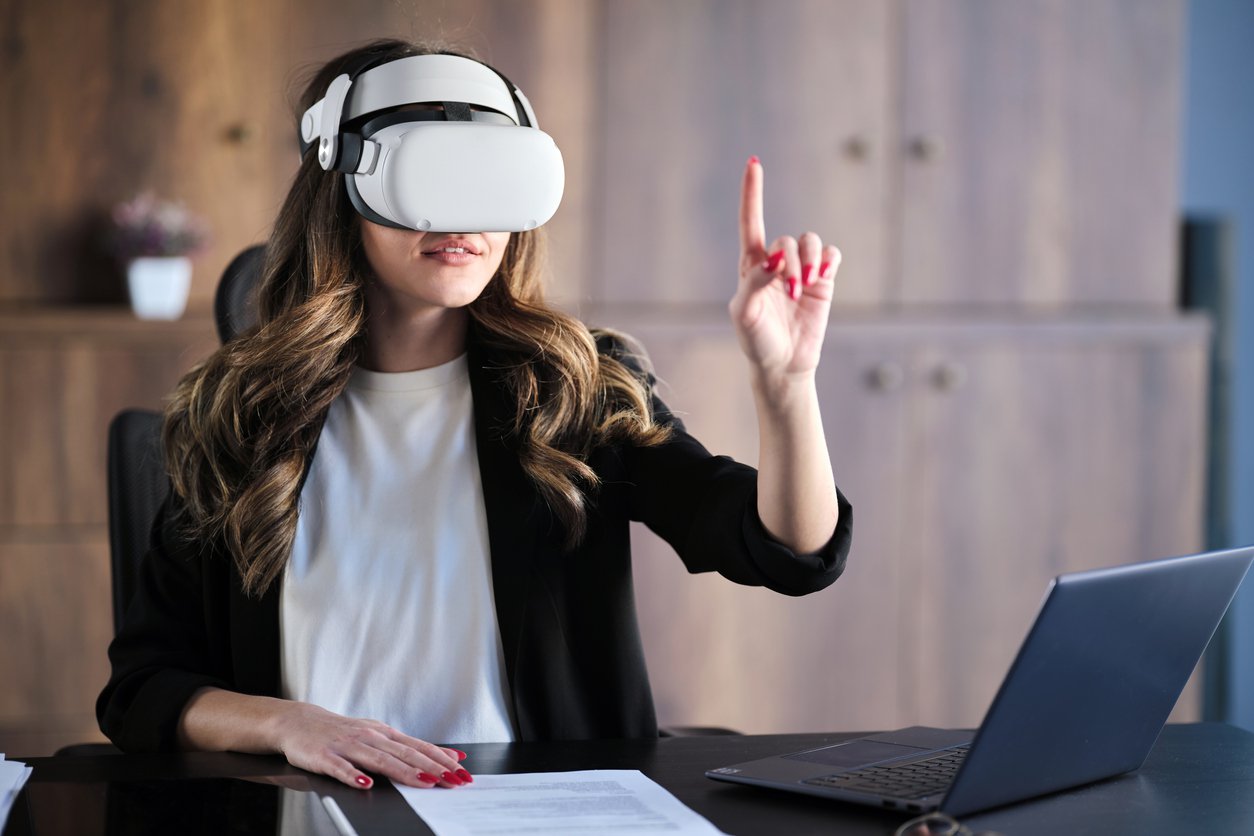 A woman in a white shirt and black jacket sites at her desk, her laptop open in front of her and one hand raised in front of her, as she uses a VR headset to access a fintech app in the Metaverse.