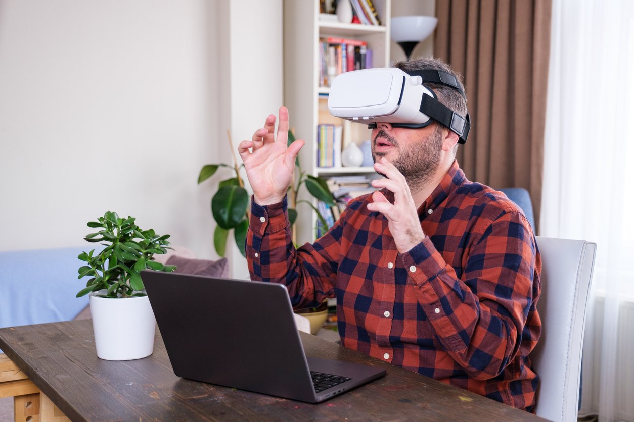 A man in a long-sleeved flannel shirt sits comfortably at his desk at home, using a VR headset to access the Metaverse.