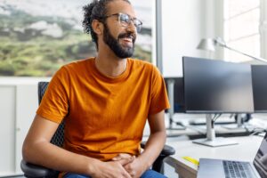A smiling digital engineer in a bright orange shirt sits as a desk in a brightly lit office in this representation of intelligence engineering.
