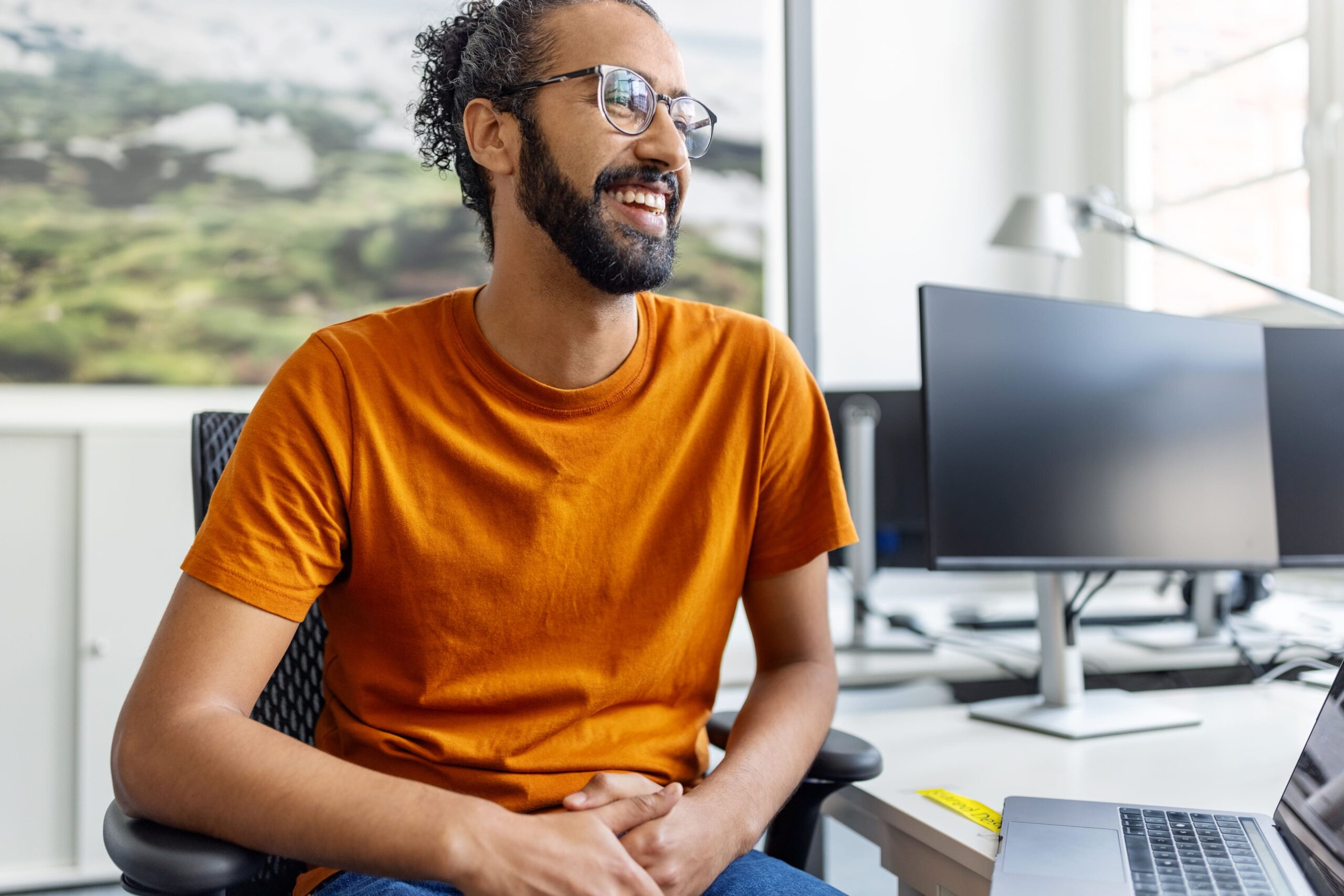 A smiling digital engineer in a bright orange shirt sits as a desk in a brightly lit office in this representation of intelligence engineering.