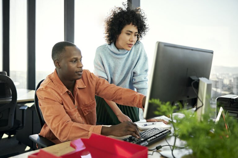Two diverse financial technology professionals collaborating at a modern office workstation, reviewing code on a desktop screen—representing engineering-led innovation in financial services.