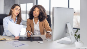 Two multiethnic businesswomen collaborating at a computer in a modern office, representing the shift toward embedded finance, ecosystem thinking, and intelligent value creation in financial services.