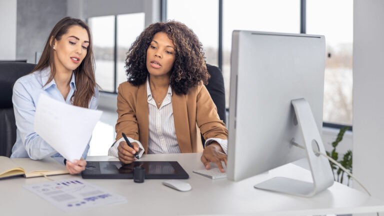Two multiethnic businesswomen collaborating at a computer in a modern office, representing the shift toward embedded finance, ecosystem thinking, and intelligent value creation in financial services.