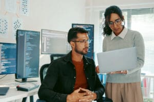 A man and a woman in an office setting. The man is sitting in a chair looking at the woman. The woman is standing and holding a laptop, showing something on the screen to the man. In the background are several computer monitors displaying code and charts on a wall.