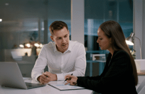 Two business professionals in a modern office are collaborating over printed analytics and a laptop during an evening strategy meeting, discussing data-driven planning and responsible AI in financial services while reviewing key insights together.
