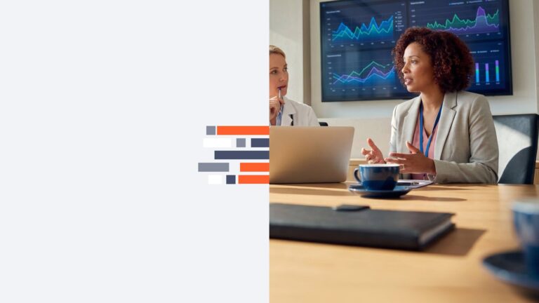 Two businesswomen in a meeting with a laptop, discussing charts displayed on a large monitor behind them, in a boardroom setting.