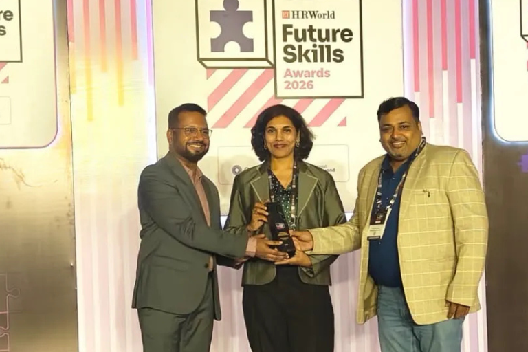 Three award recipients on a stage at the HRWorld Future Skills Awards 2026 ceremony; a man on the left helps a woman in the center hold a trophy while another man stands on the right.