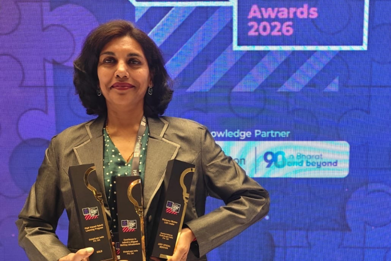 Woman in a gray blazer posing with two black and gold award trophies at an awards ceremony, standing in front of a blue stage backdrop that says 'Awards 2026'
