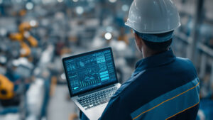 Industrial worker in hard hat using a laptop to monitor factory data and screens in the background