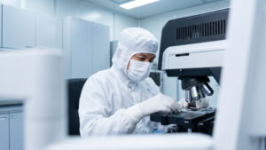 Hero-Reliable-AI-Case-Study - GlobalLogic Technician in an orange lab jacket operating semiconductor inspection equipment, using a touchscreen control panel beside a precision microscope and wafer processing unit in a clean, high-tech lab.