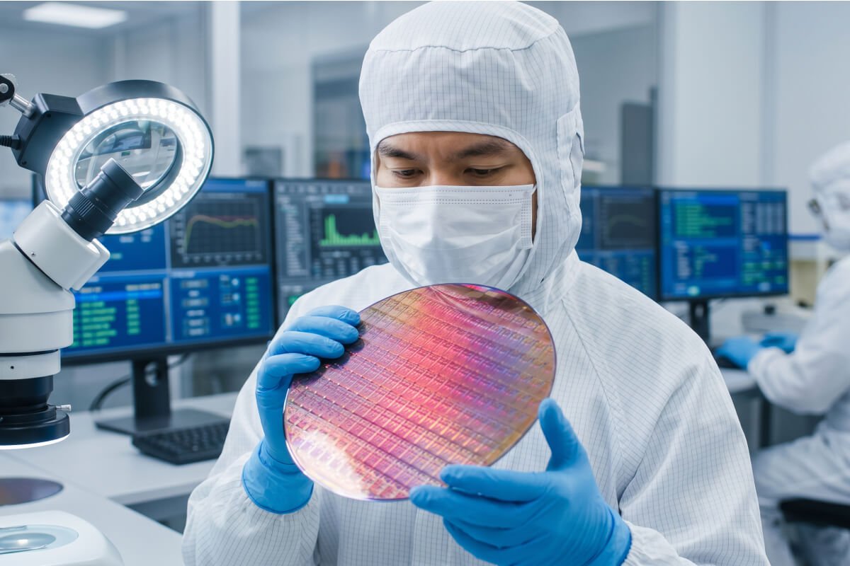 Reliable-AI-Case-Study-Image2 - GlobalLogic Scientist in a cleanroom suit and mask holds a silicon wafer near a microscope in a high-tech lab with monitors in the background