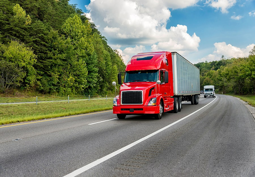 Red semi-truck driving on a two-lane highway with green trees and blue sky above.