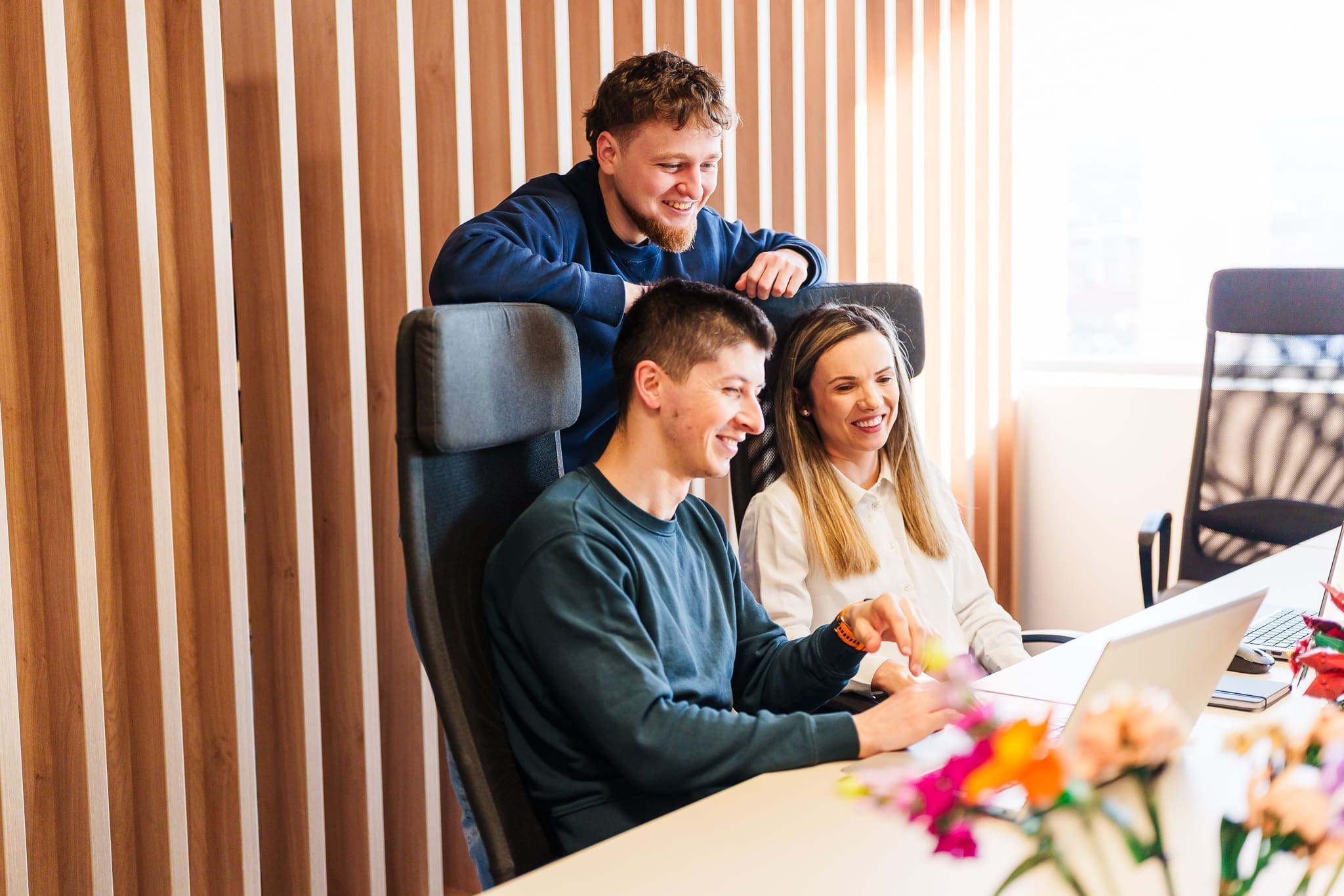 Three coworkers smile and look at a laptop at a bright office desk with a wooden slat wall in the background.