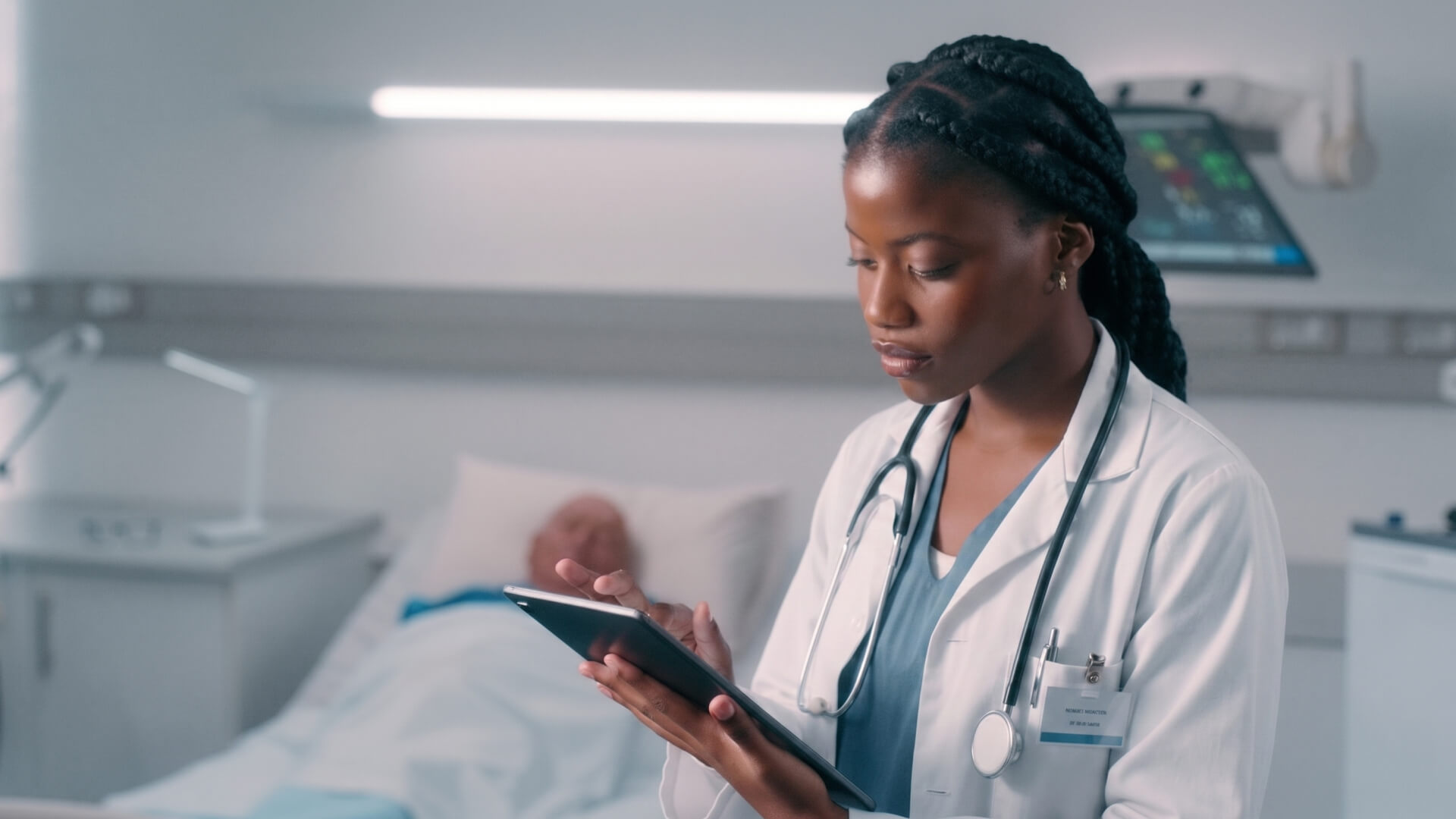 Female doctor in a white coat and stethoscope uses a tablet at the bedside in a hospital room