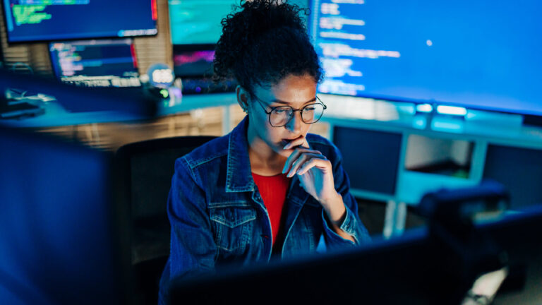 Woman with glasses in a denim jacket at a computer console, thinking with screens glowing blue around her.