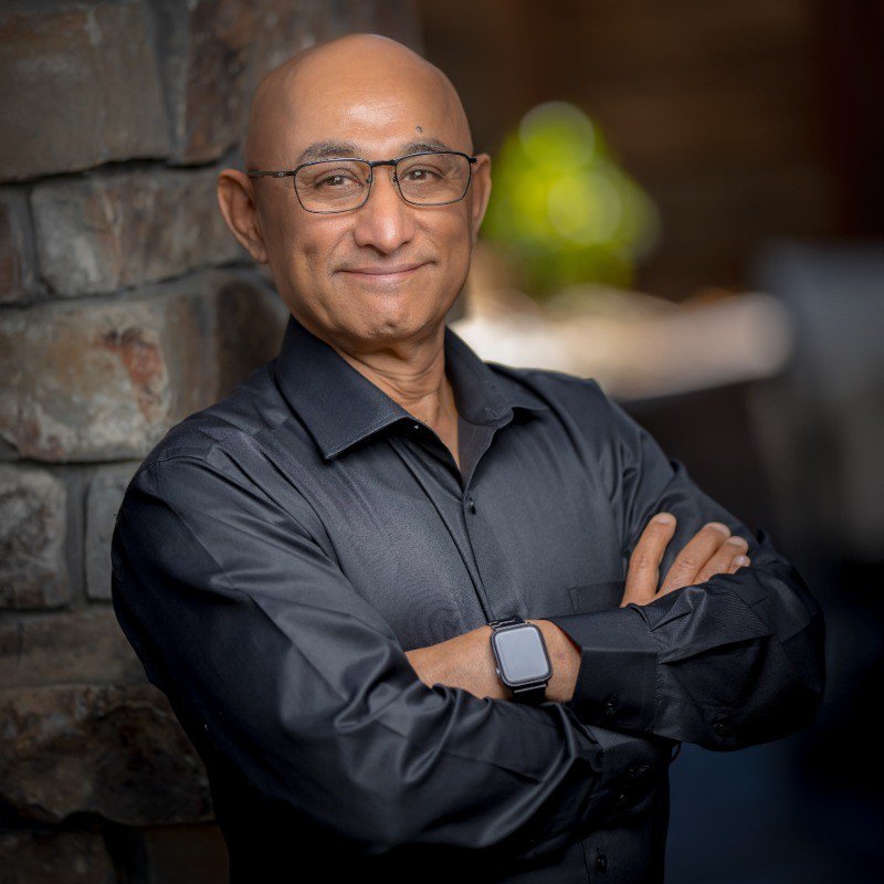 Professional portrait of a bald man with glasses, arms crossed, leaning against a stone wall in a black shirt.