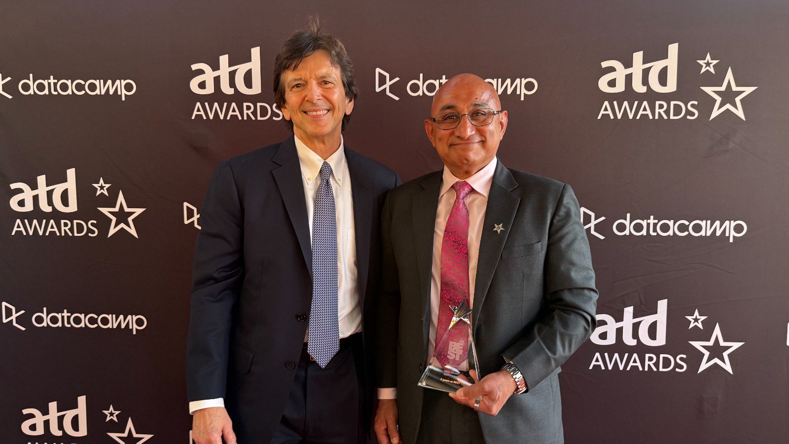 Two men in suits pose at an ATD Awards backdrop; the man on the right holds a crystal star trophy.