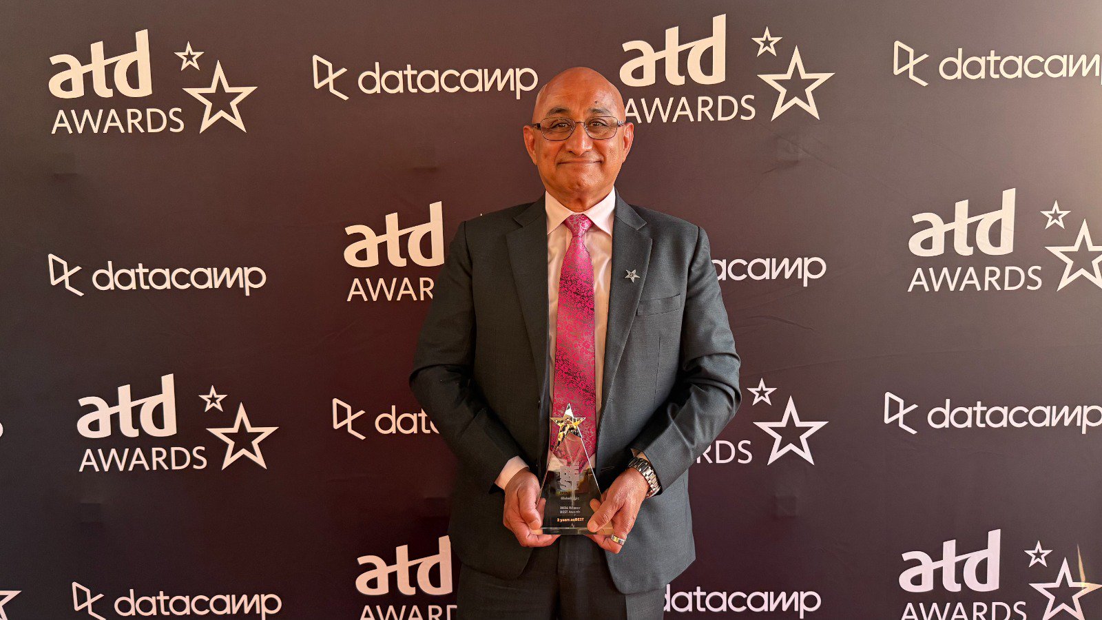 Man in a dark suit and pink tie holding a star-shaped award in front of a Datacamp/ATD Awards backdrop, smiling at the camera.