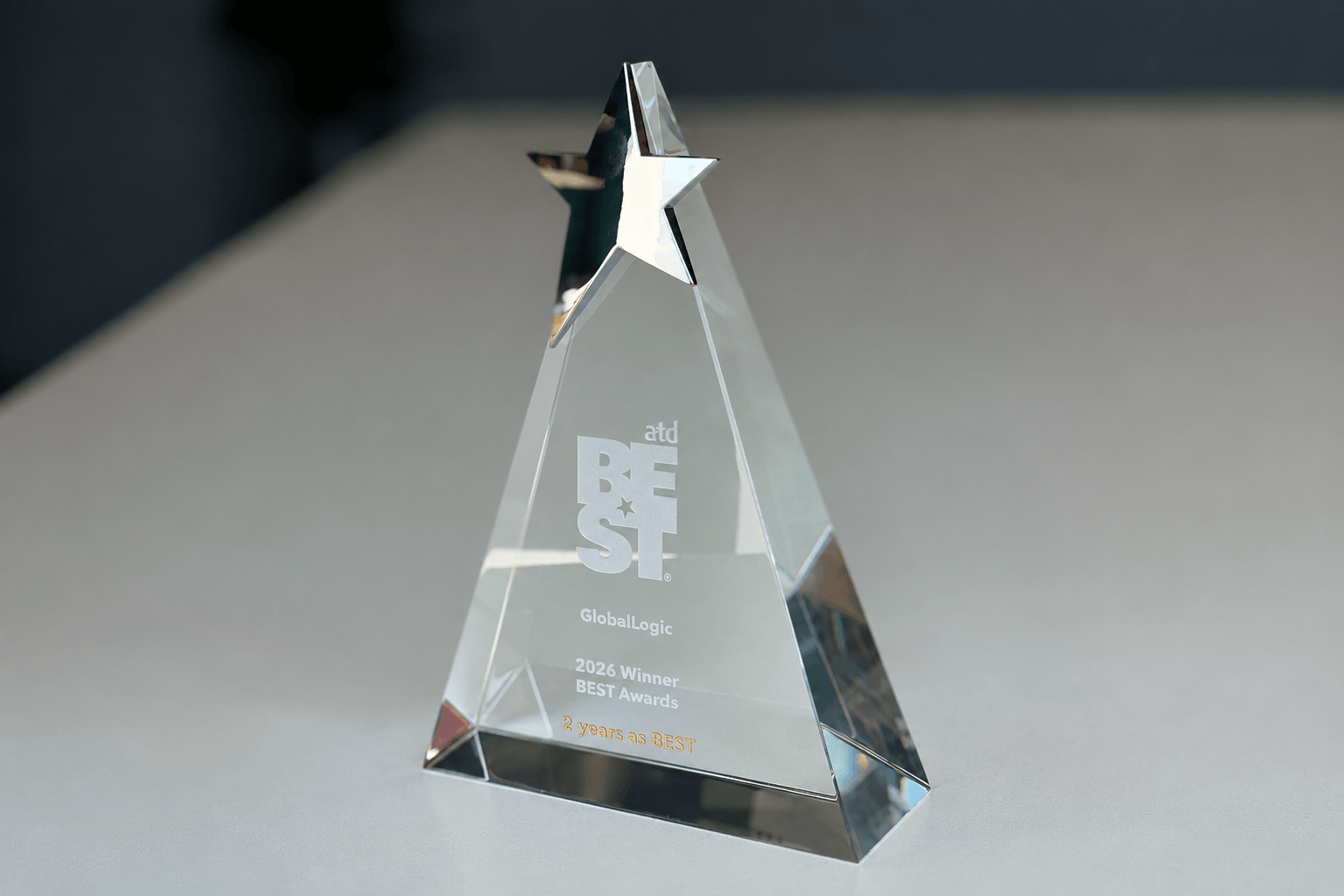 Crystal pyramid trophy with 'BEST' inscription and logos, awarded by GlobalLogic for 2026 BEST Awards on a light desk surface.