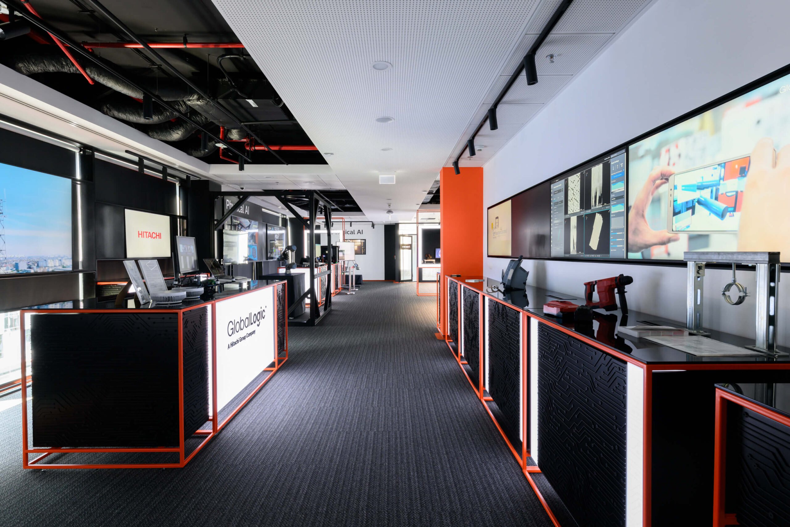 Modern tech showroom or exhibition space with large video screens, orange accents, and an exposed ceiling. The area feels like a corporate demo floor for digital techs.