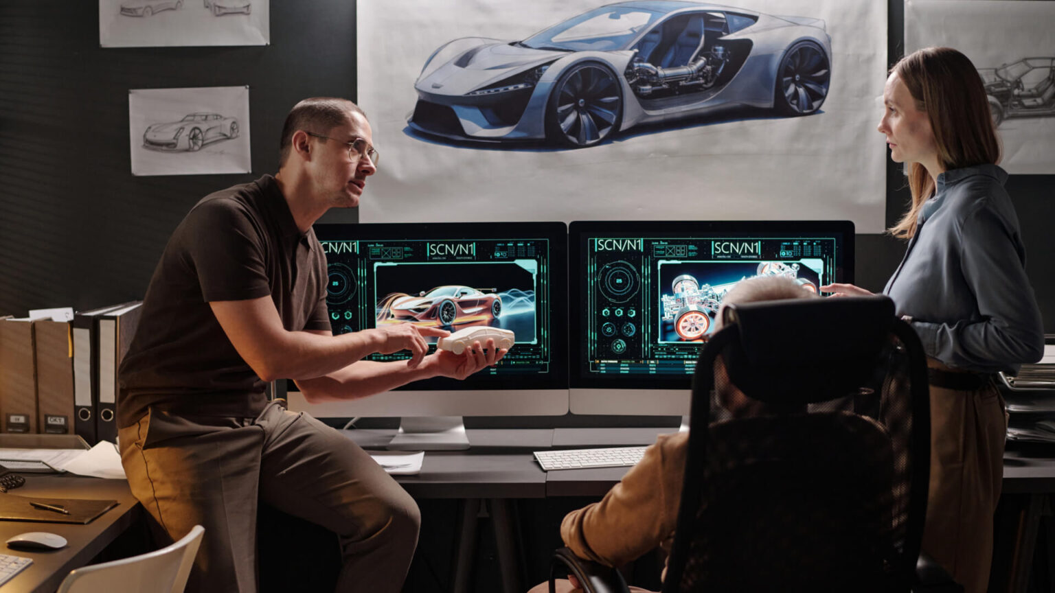 Two designers in a modern studio review a car prototype on dual computer screens, with a blue sports car diagram on the wall behind them.