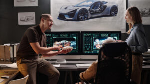 Two designers in a modern studio review a car prototype on dual computer screens, with a blue sports car diagram on the wall behind them.