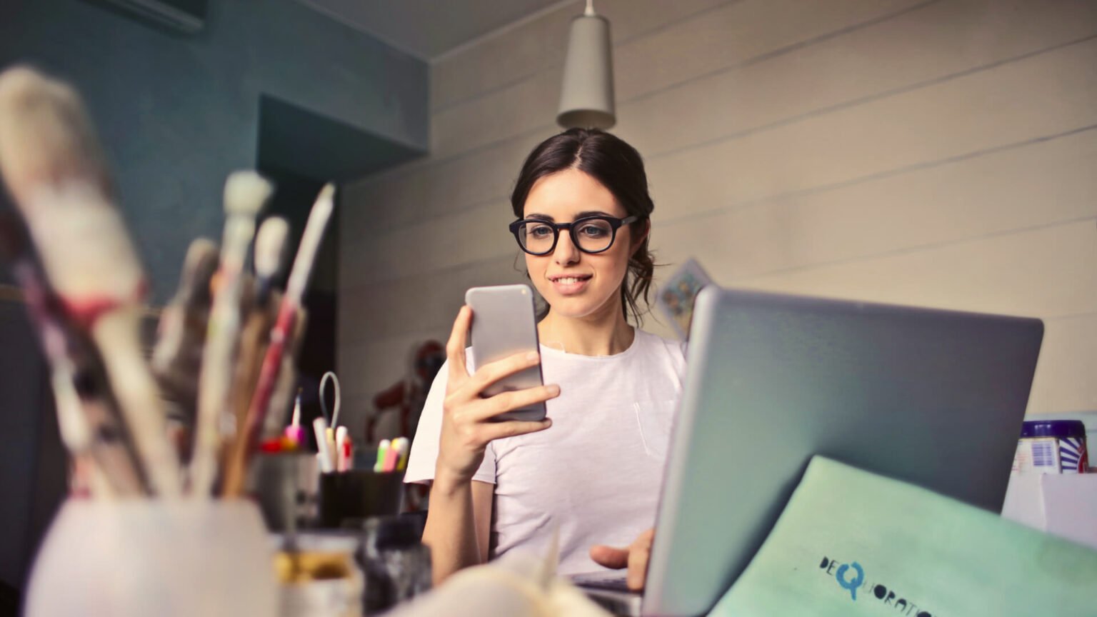 Woman with glasses sits at a desk, looking at her smartphone beside a laptop in a studio.