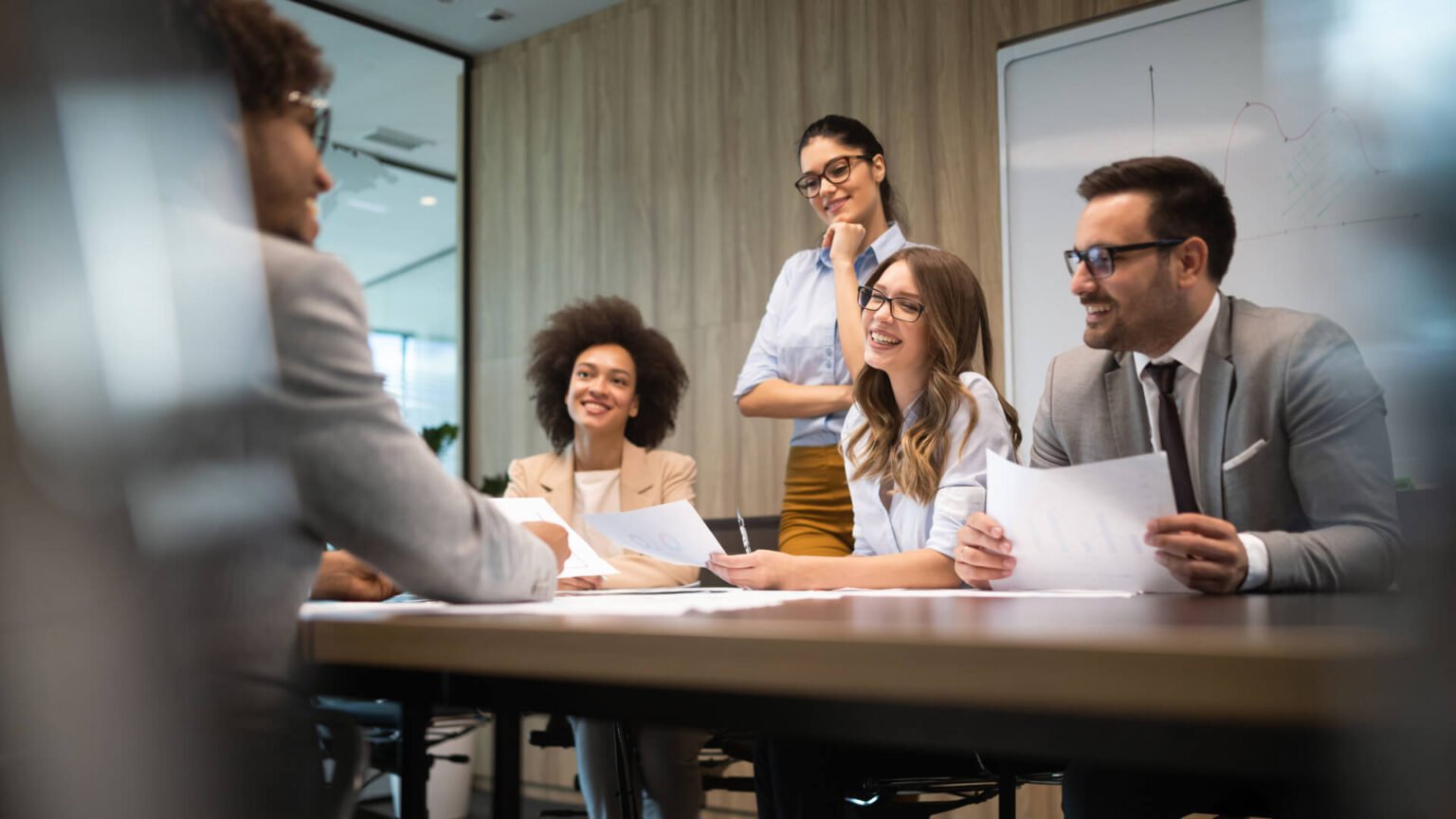 Group of five professionals in a meeting, smiling and discussing papers around a conference table in a modern office.