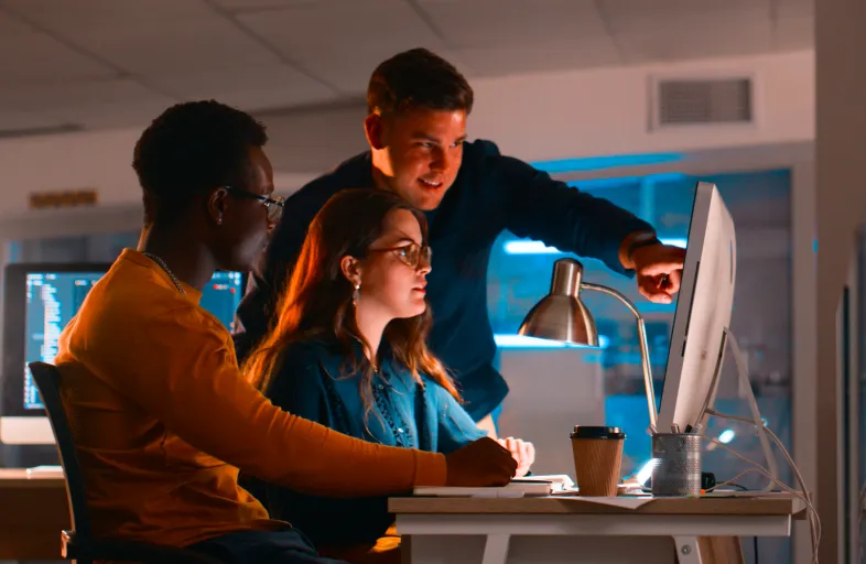 Three colleagues collaborate at a computer in a dimly lit office, one pointing at the monitor as others look on.