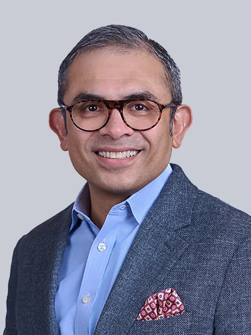 Professional headshot of a smiling man with glasses, wearing a blue shirt and gray blazer with a red patterned pocket square.