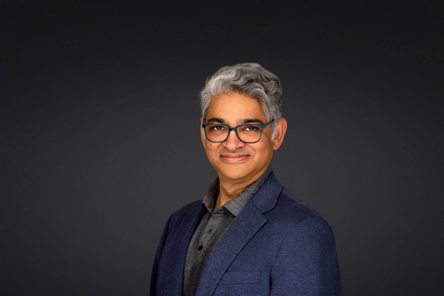 Portrait of a middle-aged man with gray hair and blue-framed glasses, smiling, in a navy blazer against a dark backdrop.